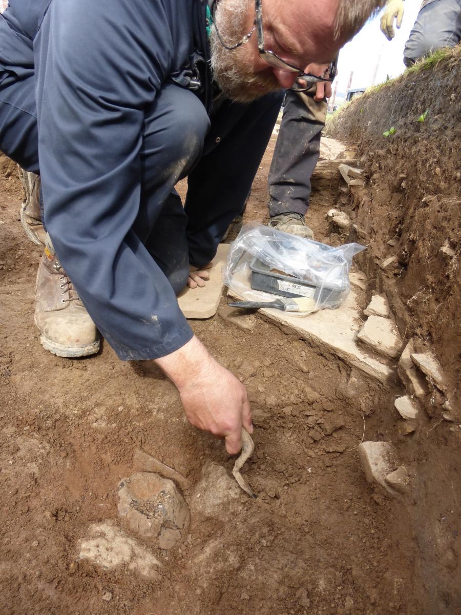 A photograph showing a man in a trench excavating pottery sherds with a trowel